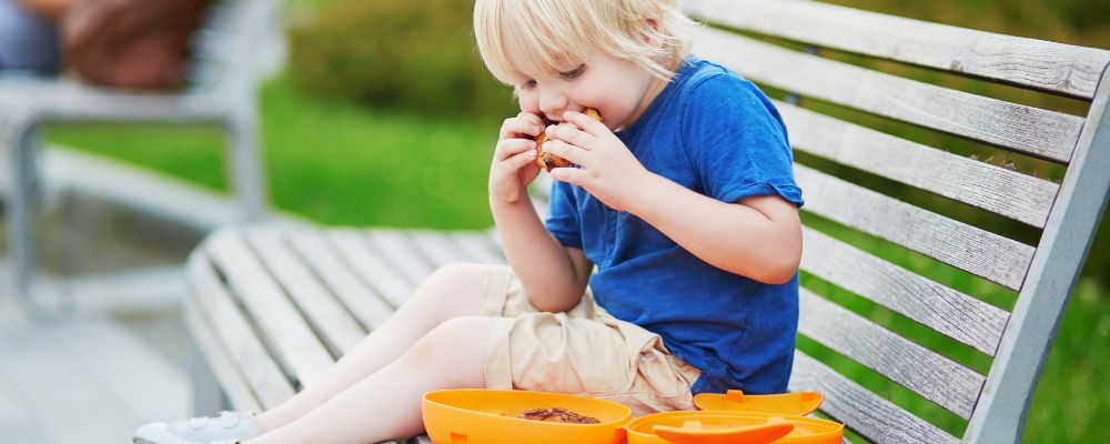 A toddler sitts ona bench while eating a sandwich from a lunchbox