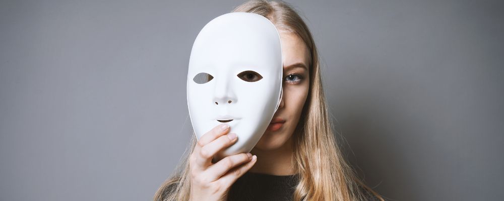 The face of a young blonde woman emerges from behind a white mask